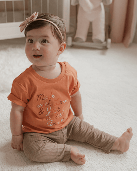 A baby girl smiling with a gold headband and a shirt that says "made with love and just a little anxiety."