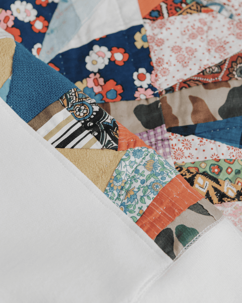 A natural cotton crewneck hanging from a hanger with vibrant orange and blue quilted sides and a quilt on the bottom of the frame.