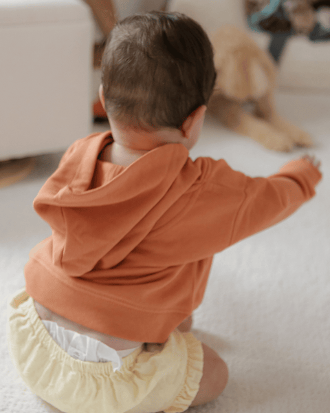 A baby smiling in a yellow diaper and a pumpkin-colored organic cotton hoodie.