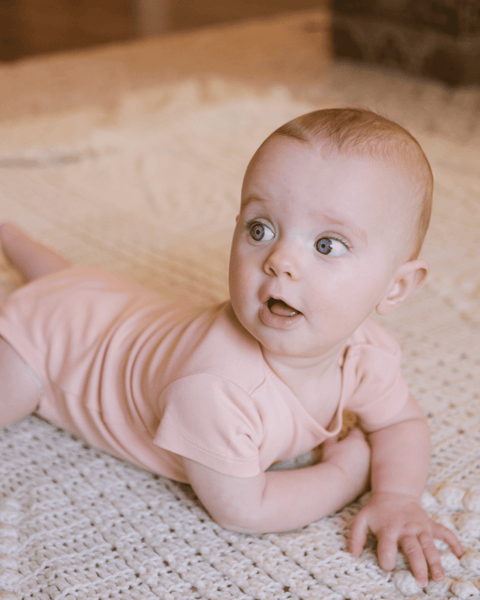 A smiling baby girl in two photos, one wearing the apricot bubuleh onesie and one in the butter onesie, both made with organic cotton. She's on a vintage white blanket for tummy time.