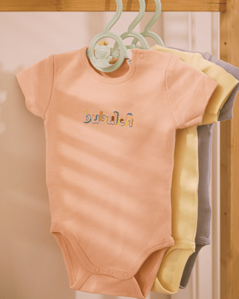 The apricot color baby onesie by bubuleh hanging on a wooden rack.