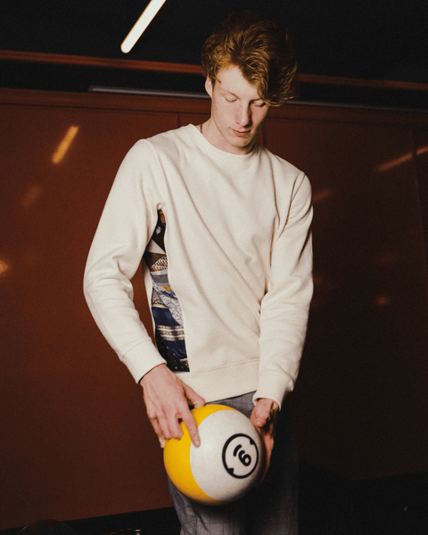 A man stands holding a yellow bowling ball and shows off the sides of his white sweater which is made with quilted ties.