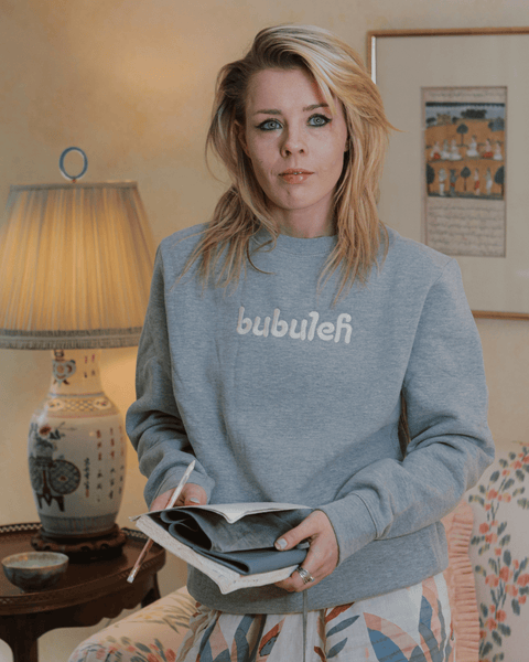 A girl smiles off camera with a journal in her hands, wearing a unisex handcrafted bubuleh crewneck.