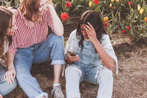 A photo of three girls sitting outside in jean overalls, schmoozing, while one looks at her phone. There are red and yellow roses behind them.