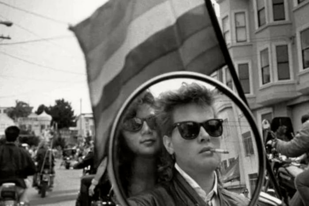 A photo of two women on motorcycles holding a gay pride flag in the Castro in 1988. Photo is black and white.