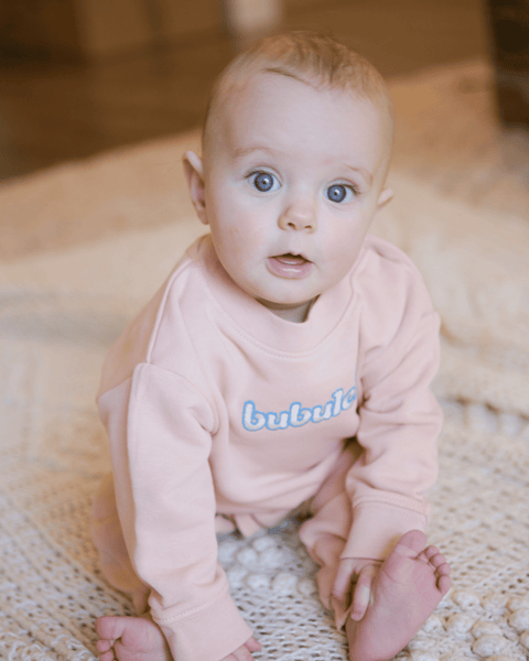 A baby wearing an apricot-colored crewneck while smiling. The word "bubuleh" is embroidered on the front.
