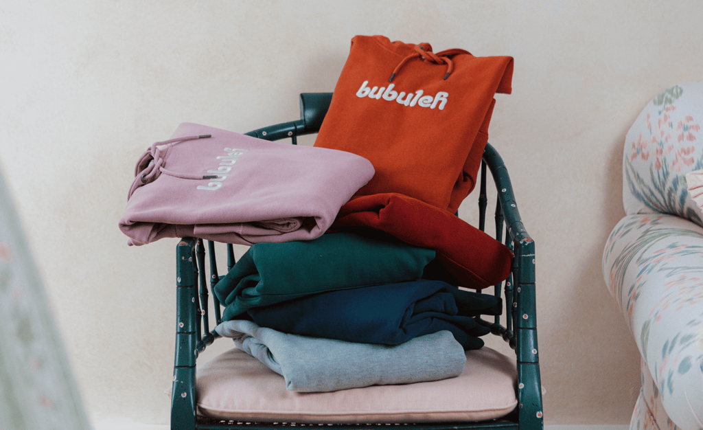 A photo of 6 bubuleh hoodies, all different colors, on a vintage green chair in a grandma's floral-inspired fall living room.
