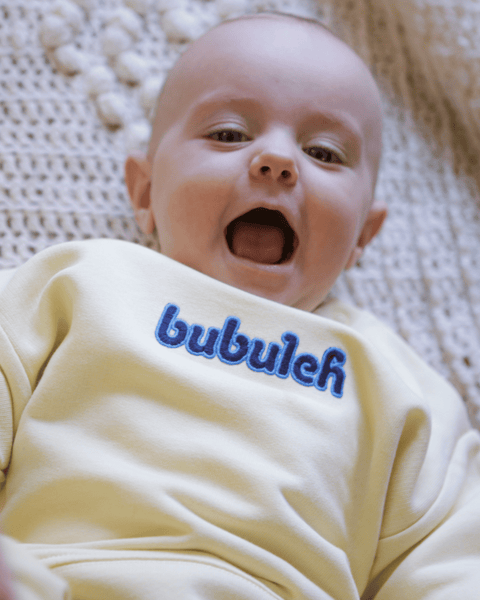 A baby girl laughing with her Mom on the floor wearing a crewneck that says "bubuleh" on the front. Bubuleh means sweetheart in Yiddish.