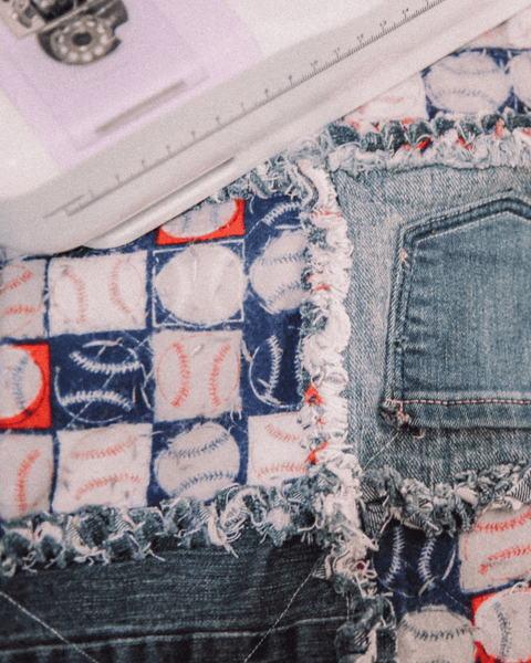 A denim and baseball-print vintage quilt, handmade with the seams up, next to a pink and purple sewing machine. The image is overexposed showing the details of the red and blue quilt.