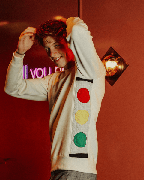 A red-headed model smiling and hugging himself while wearing the bubuleh joyride quilted sweater, with the words "will you perform for me" in the background in neon lights.