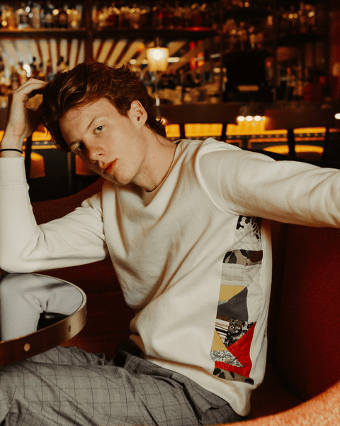 A young model posing in a retro bar in an orange chair wearing a white upcycled sweater with red and yellow patches on the sides.