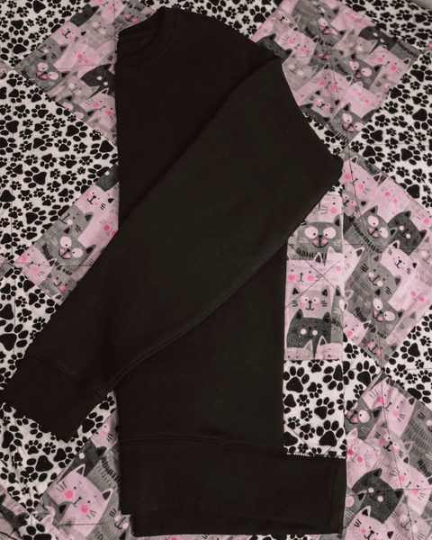 A photo of a vintage black and pink quilts with patchwork of dog paws and cat faces, with a black crewneck folded on top.