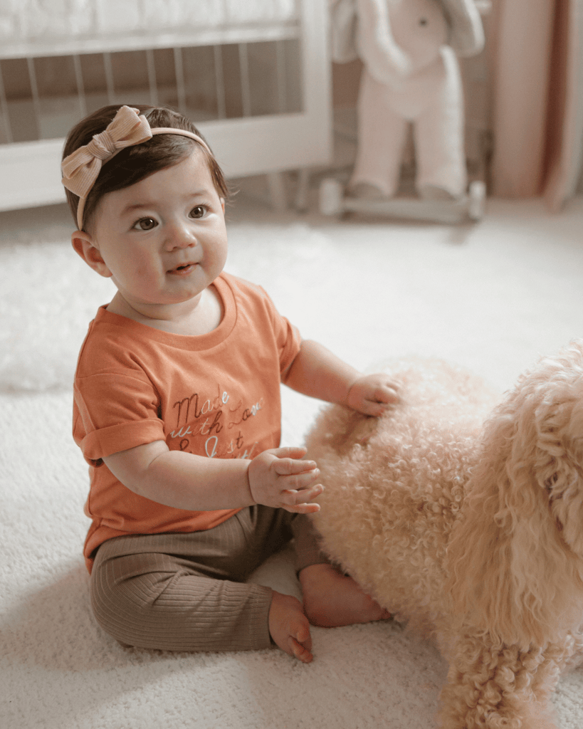 A baby girl smiling with a gold headband and a shirt that says "made with love and just a little anxiety" with her golden dog next to her on the floor.