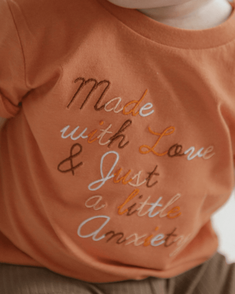 A detail shot of 4 different embroidery threads on a baby shirt saying "made with love and just a little anxiety"