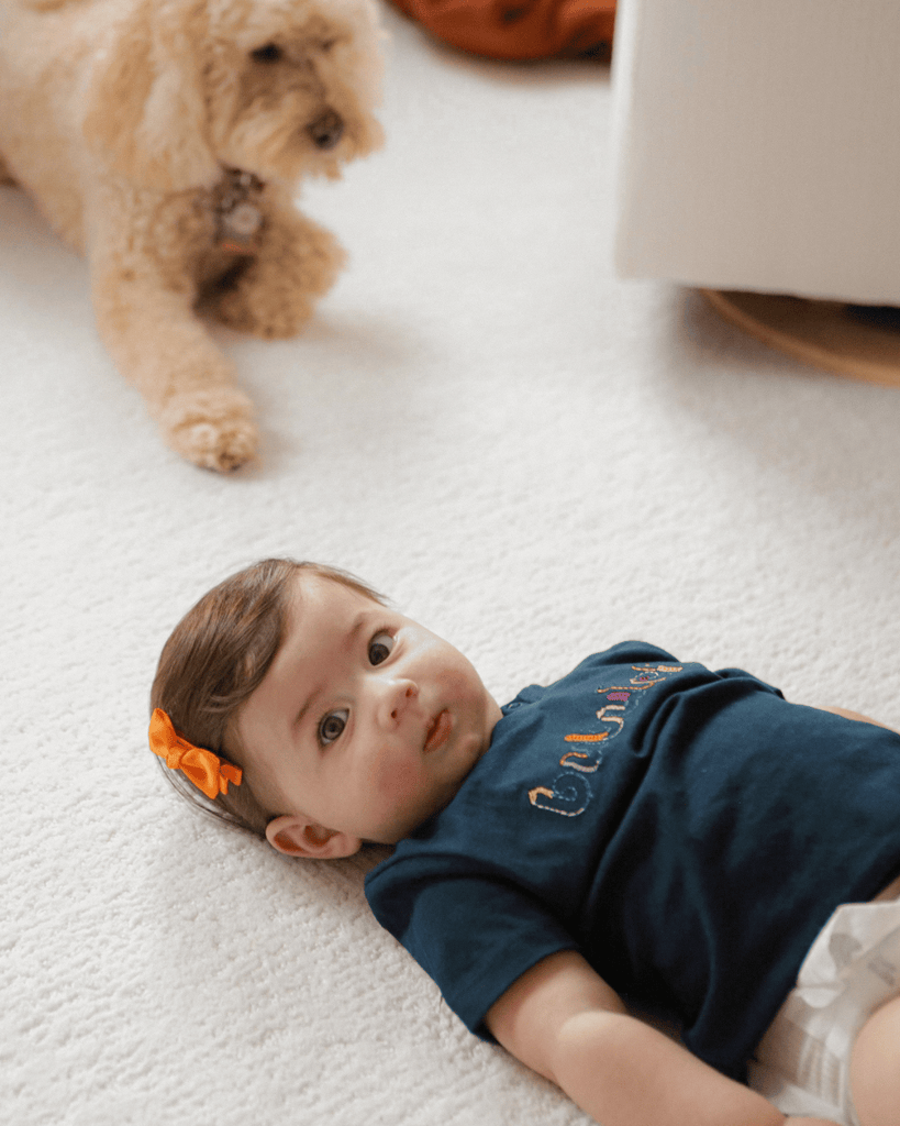 A baby girl and her dog playing on the floor in a diaper and blue bubuleh mini t-shirt.