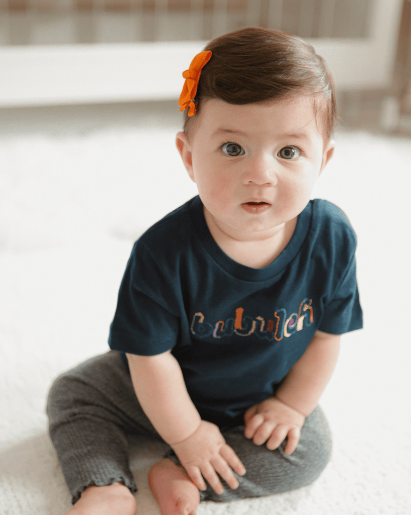 A baby girl in a navy blue shirt that says "bubuleh" in 6-color embroidery.