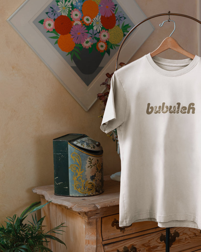 A photo of an off-white t-shirt (made in portugal) on a valet stand with a floral painting and vintage chest in the background, where the shirt says bubuleh on the front in two different fonts.