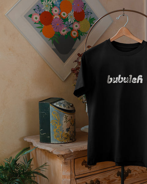 A photo of a black t-shirt on a valet stand with a floral painting and vintage chest in the background, where the shirt says bubuleh on the front in two different fonts.