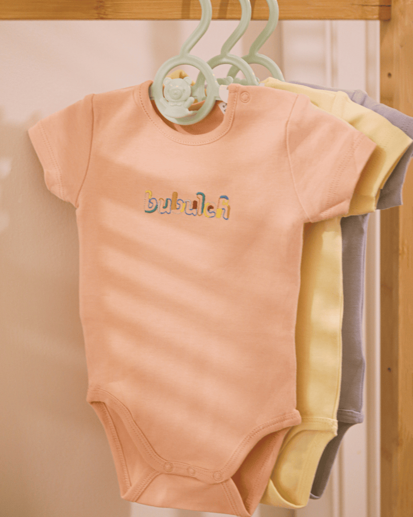 The apricot color baby onesie by bubuleh hanging on a wooden rack.