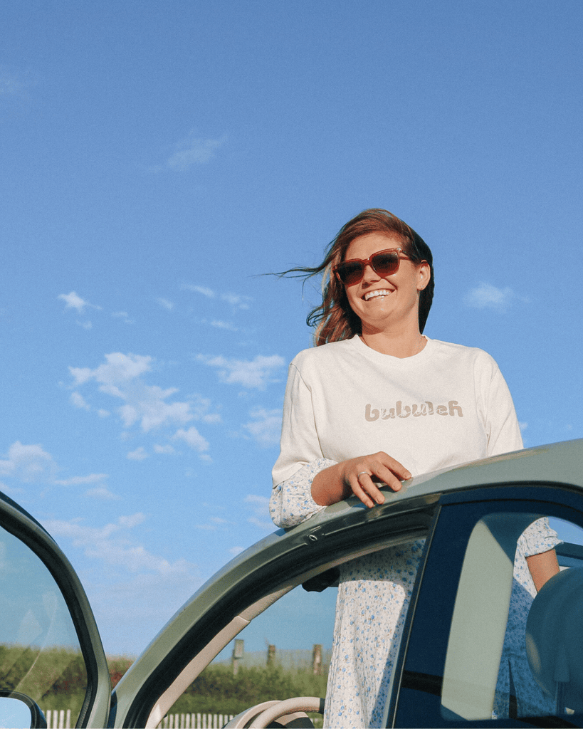 A girl is smiling standing through a green fiat sunroof looking off camera whiie wearing a bubuleh t-shirt.