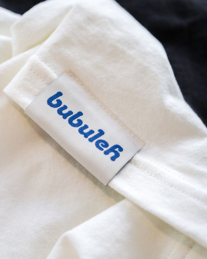 A sewn-on bubuleh tag in bright blue on a white tank top against a black tank top in the background.