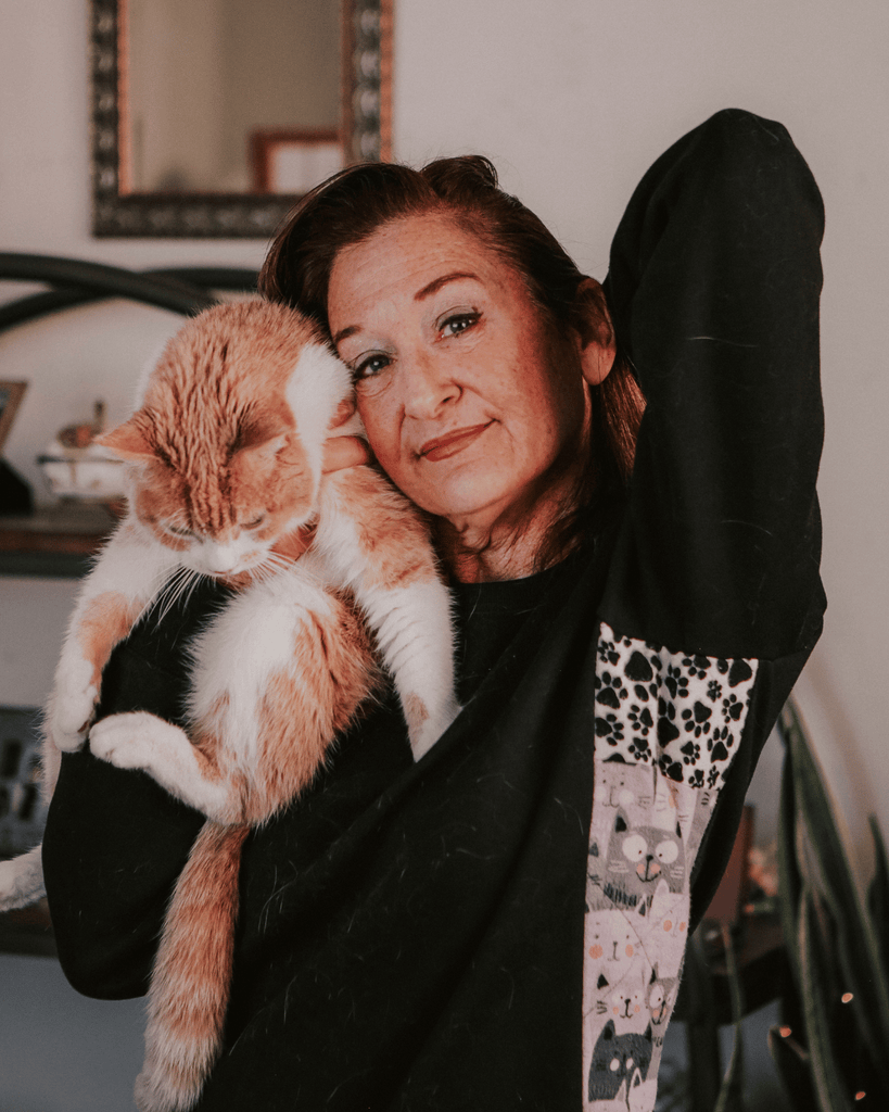 A woman in cat-themed sweater holds up her cat like Simba in her Los Angeles apartment. There's a vintage photo of a dog in the background.