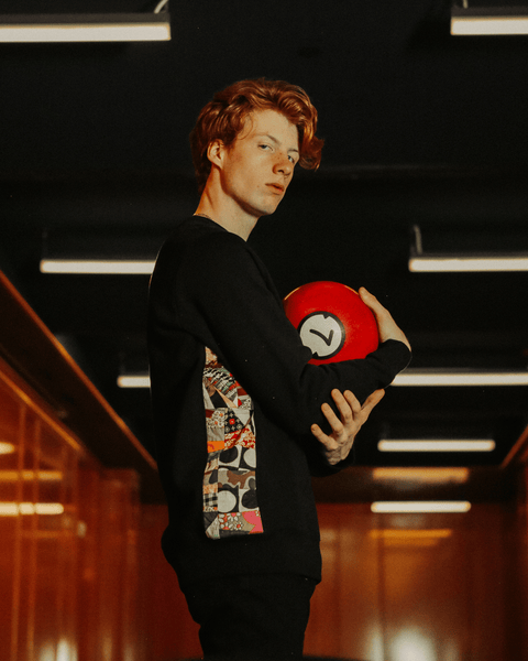 A red-haired young man holding a red #7 bowling ball in the bubuleh sweet but psycho crewneck which has all different colors and patterns on the sides. He's looking straight at the camera.