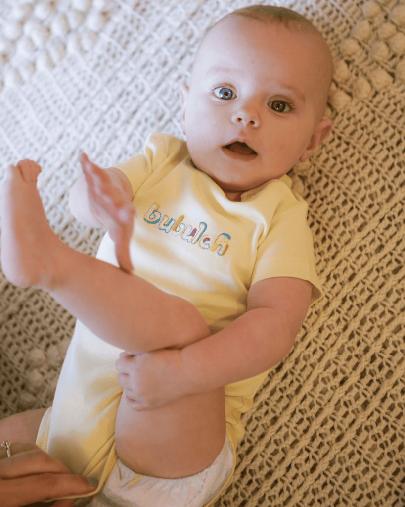 A baby girl wearing the organic cotton bubuleh onesie in butter, grabbing her leg and looking up at the camera smiling.