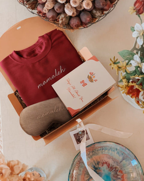 An aerial photo of bubuleh's mamaleh gift box for mother's day, which shows a maroon shirt, a linen sleep mask a and a Mother's Day card besides flowers.