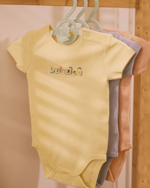 The butter color baby onesie by bubuleh hanging on a wooden rack.