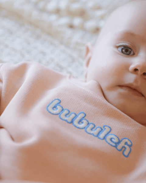 An adorable close-up of a baby wearing an apricot shirt that says bubuleh on the front.