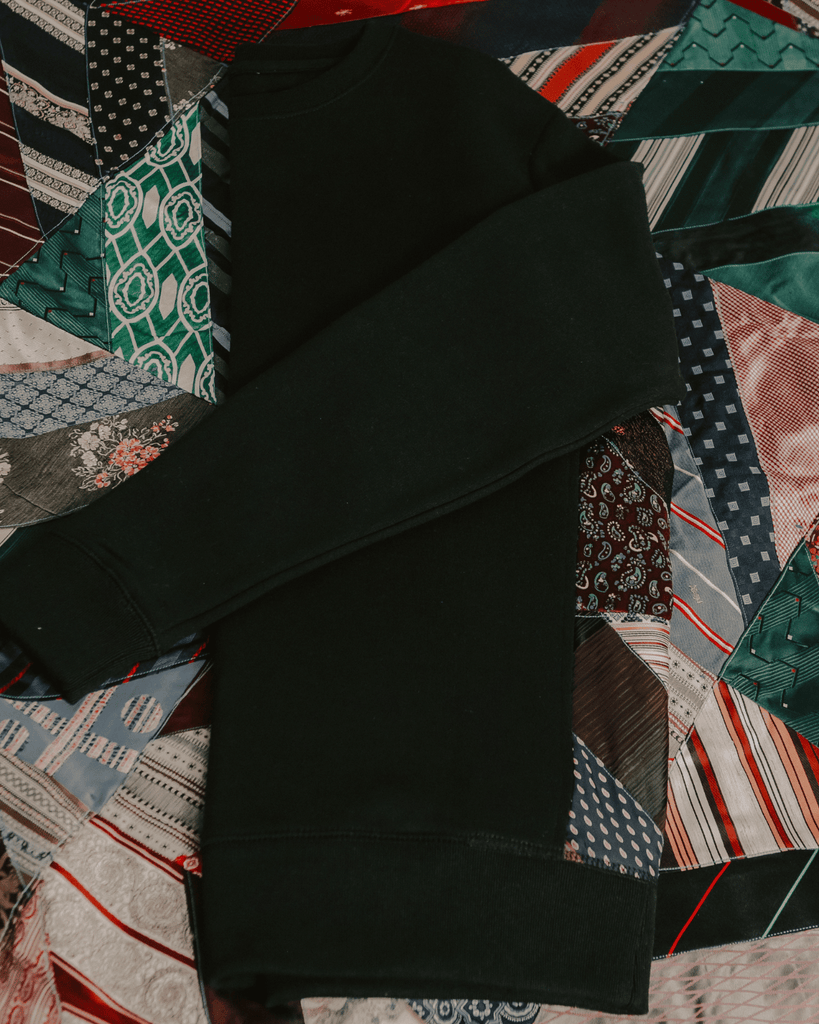 A flat lay photo of a black sweater against a quilt made of ties.