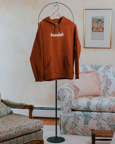 A cinnamon colored french terry organic cotton bubuleh hoodie hanging on a circular valet stand with a floral couch in the background.