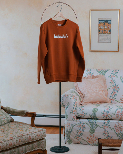 A handcrafted cinnamon crewneck on a circular valet stand in a bubbe / grandma's living room.