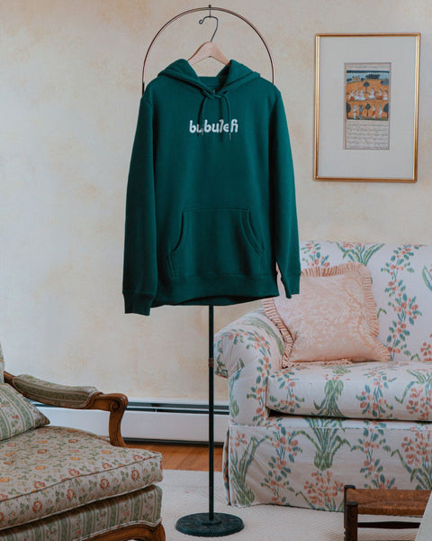A green organic cotton bubuleh hoodie hanging on a circular valet stand with a floral couch in the background.