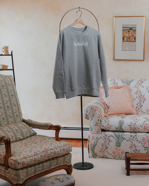 A handcrafted heather gray crewneck on a circular valet stand in a bubbe / grandma's living room.
