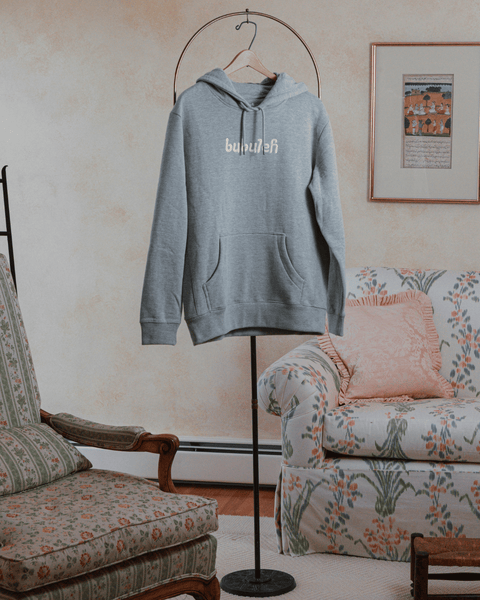 A heather gray french terry organic cotton bubuleh hoodie hanging on a circular valet stand with a floral couch in the background.