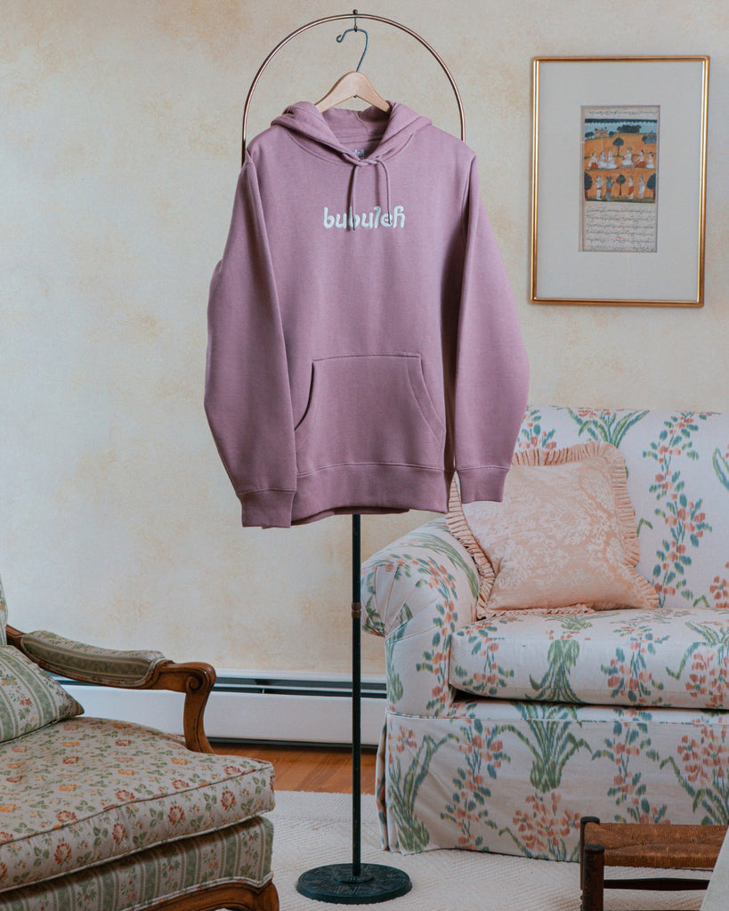 A lavender bubuleh hoodie hanging on a circular valet stand with a floral couch in the background.
