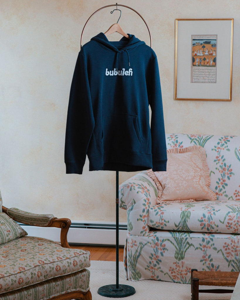 A navy blue french terry organic cotton bubuleh hoodie hanging on a circular valet stand with a floral couch in the background.