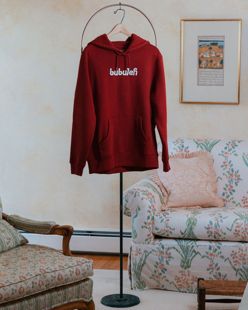 A red french terry organic cotton bubuleh hoodie hanging on a circular valet stand with a floral couch in the background.