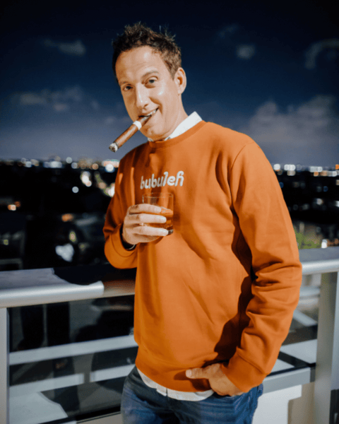 Comedian Elon Gold smokes a cigar with a glass of whiskey in his hand while wearing a bubuleh cinnamon crewneck.