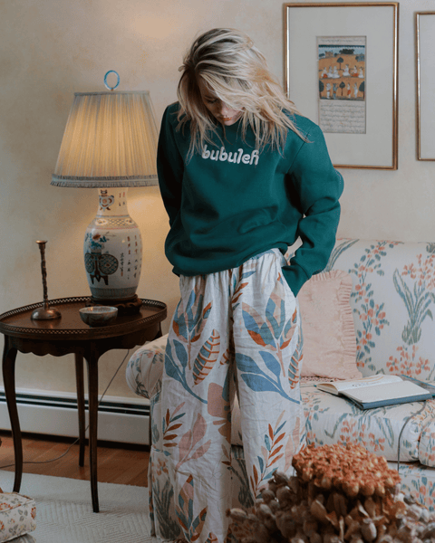 A woman wears a green bubuleh crewneck in a Grandma's living room, with dry flowers in the foreground and a floral couch in the background.