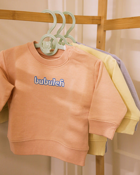 A clothing rack with three crewnecks for babies, with the one in front being apricot. They all say "bubuleh" embroidered in the front.