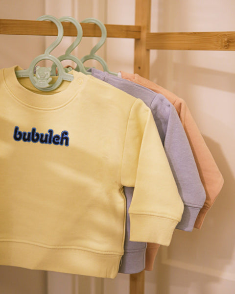 A clothing rack with three crewnecks for babies, with the one in front being butter. They all say "bubuleh" embroidered in the front.
