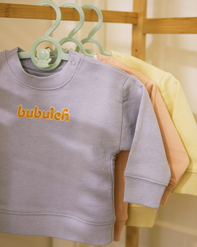 A clothing rack with three crewnecks for babies, with the one in front being blueberry blue. They all say "bubuleh" embroidered in the front.