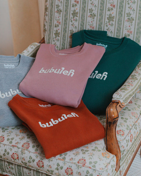 A pile of bubuleh handcrafted crewnecks in four colors on a vintage floral chair.
