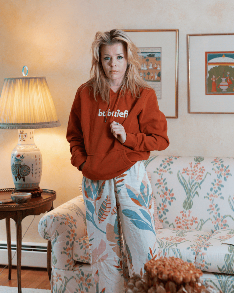 A bubuleh model looks directly into the camera while wearing a cinnamon colored organic cotton french terry hoodie inspired by the grandma's living room that the photo is taken in.