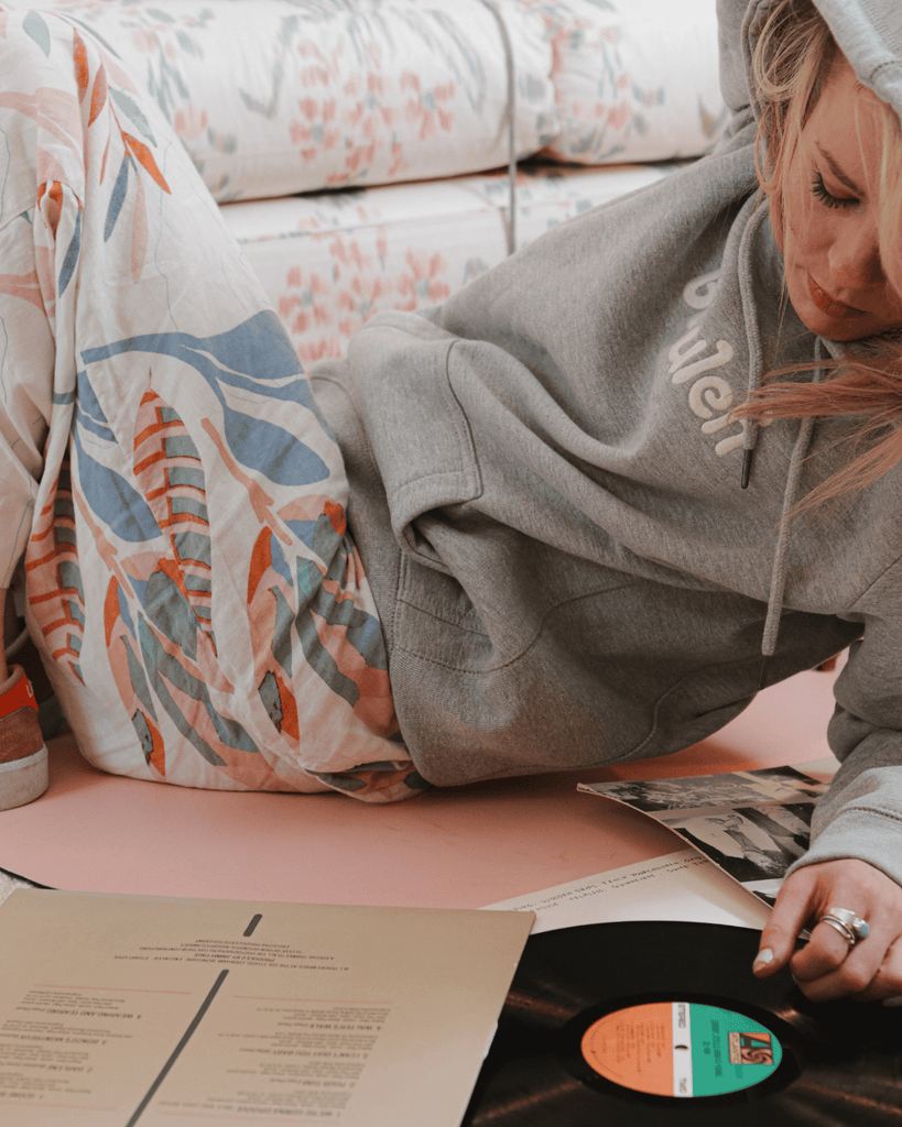 A blonde bubuleh model poses in a bubuleh hoodie while looking at a retro Stevie Nicks album and journaling.