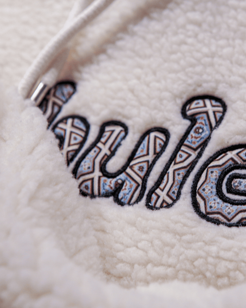 A closeup details shot of the sherpa hoodie metal drawstrings and raised neutral-tone embroidery.
