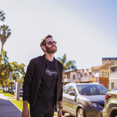 A lifestyle shot of a man in sunglasses wearing a black faygeleh tee and a black jacket.